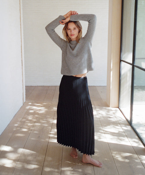 A woman stands barefoot in a bright, sunlit room with light wood floors and white walls, wearing a gray long-sleeve knit sweater and a long black pleated skirt.