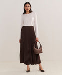 A woman poses against a plain light background, wearing a long-sleeved white top, the Pleated Skirt in chocolate, brown heels, and holding a small brown handbag.