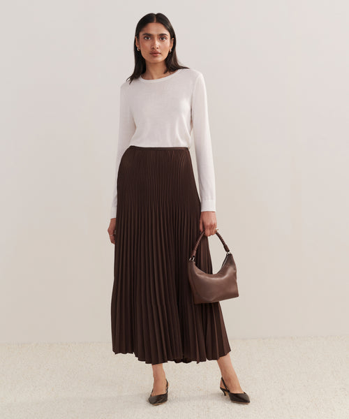 A woman poses against a plain light background, wearing a long-sleeved white top, the Pleated Skirt in chocolate, brown heels, and holding a small brown handbag.