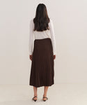 A woman with long dark hair, dressed in a white long-sleeve top and the Pleated Skirt, stands facing away from the camera against a plain light background.