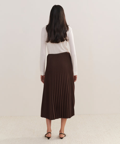 A woman with long dark hair, dressed in a white long-sleeve top and the Pleated Skirt, stands facing away from the camera against a plain light background.