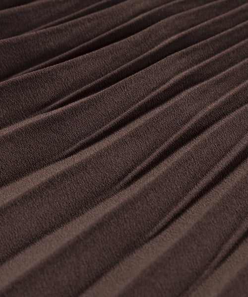 Close-up of the Pleated Skirt in brown, showing detailed texture and diagonal pleats that create raised and recessed lines across the fabric.