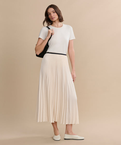 A woman stands against a beige background wearing a short-sleeved white top, the Pleated Skirt in ivory, white flats, and carrying a black shoulder bag.
