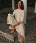 A woman wearing a cream ribbed knit sweater and matching pleated skirt sits barefoot on a weathered wooden platform outdoors.