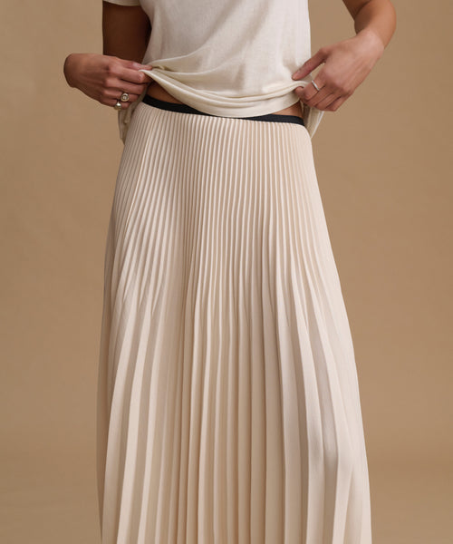 A close-up of a person wearing a cream-colored, long pleated skirt with a black waistband, paired with a matching cream top, set against a solid beige background.