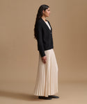 A woman stands in profile wearing a black cardigan over a white top, paired with a long cream pleated skirt and black shoes, against a plain beige studio background.