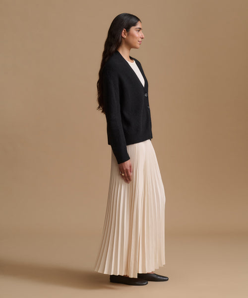 A woman stands in profile wearing a black cardigan over a white top, paired with a long cream pleated skirt and black shoes, against a plain beige studio background.