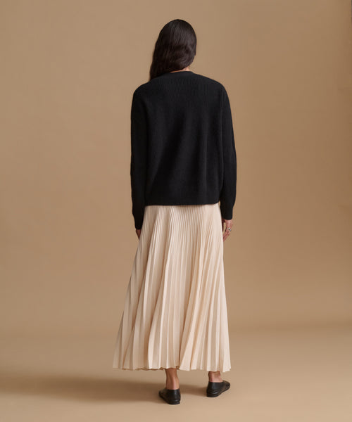 A woman is shown from behind wearing a black knit sweater and a cream pleated maxi skirt, standing against a solid beige studio background.