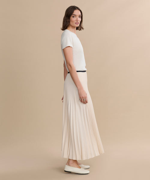 A woman with shoulder-length brown hair and a neutral expression stands sideways against a beige background, wearing a long ivory Pleated Skirt, a short-sleeve white top, and white flats.