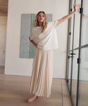 A woman stands barefoot in a minimalist room with light wood floors and white walls, wearing an oversized cream short-sleeve top and a long cream pleated maxi skirt, leaning against a glass door frame.