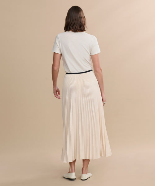 A person stands with their back to the camera, wearing a short-sleeve white t-shirt, the Pleated Skirt in ivory with a black waistband, and white flats, set against a plain beige background.