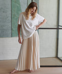 A woman stands barefoot indoors wearing an oversized white short-sleeve top and a long cream pleated maxi skirt.