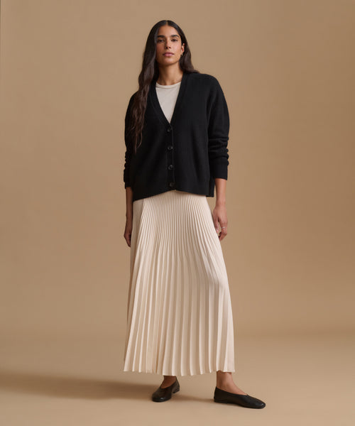 A woman stands against a warm beige studio background wearing a black button-front cardigan over a white top and a long cream pleated skirt, with black flats.