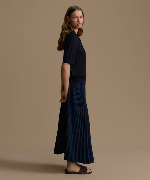 Woman wearing a navy top and navy pleated skirt against a beige background