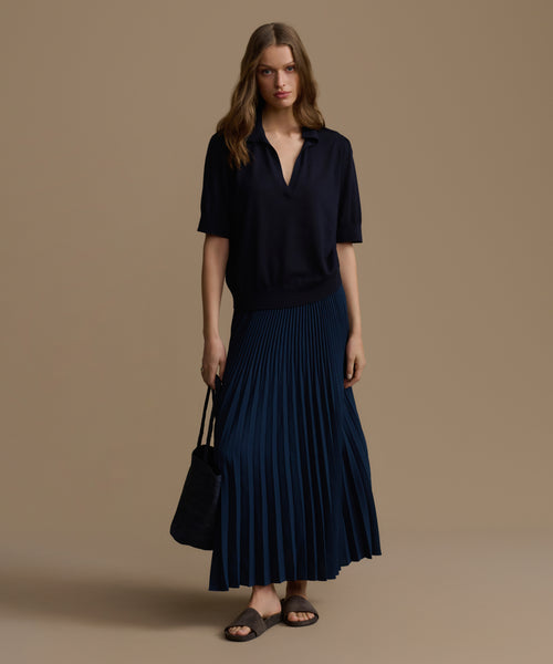 Woman wearing a navy blue top and pleated skirt holding a black handbag against a beige background