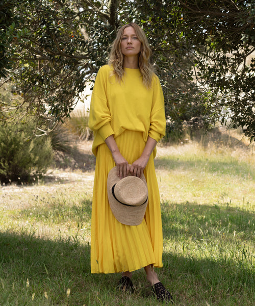 A woman stands on grass, surrounded by trees, wearing a Pleated Skirt and a loose yellow sweater. She holds a straw hat in front of her, sunlight catching the flattering drape of her Pleated Skirt.