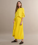 A woman in a Pleated Skirt and a loose yellow sweater stands against a plain beige background. She pairs the outfit with brown woven slip-on shoes, her long hair and neutral expression highlighting the skirts flattering drape.