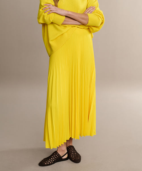 Someone wears the Pleated Skirt in bright yellow, paired with a matching sweater and black woven flats, standing arms crossed against a plain background.