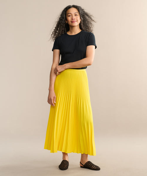 A woman with long curly hair smiles softly as she stands against a beige background, wearing a black top, black woven slip-on shoes, and the bright yellow matte Pleated Skirt with a flattering drape.