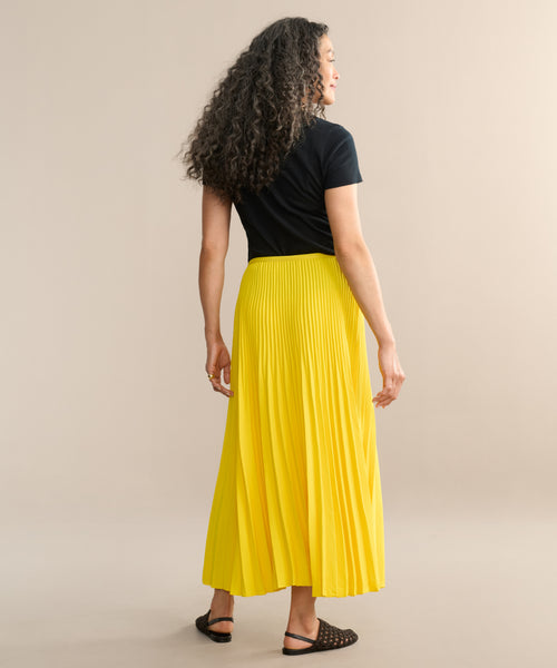 A person with curly hair, wearing a black short-sleeve top, the Pleated Skirt in bright yellow, and black sandals stands facing away against a plain beige background.