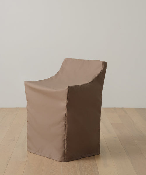 The Outdoor Montecito Dining Arm Chair Cover, in snug waterproof fabric, hides the chair’s contours with clean lines and a slightly angled back, blending seamlessly against a light wood floor and an off-white wall.