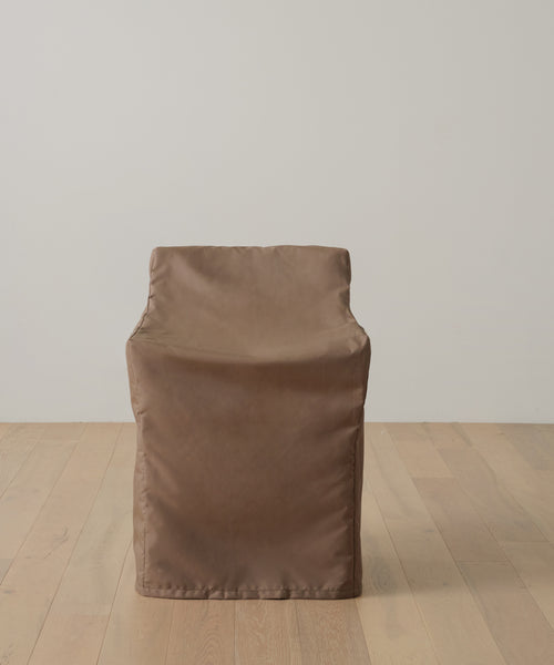 A brown, boxy armchair—shielded by the Outdoor Montecito Dining Arm Chair Cover—rests on light wood flooring against a plain white wall.