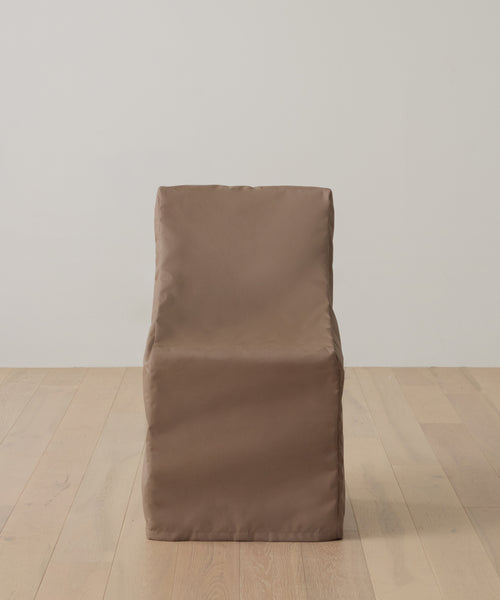 A brown, fabric-covered chair with a boxy design sits on a light wooden floor against a white wall, protected by the Outdoor Montecito Dining Chair Cover for UV resistance, lasting durability, and style.