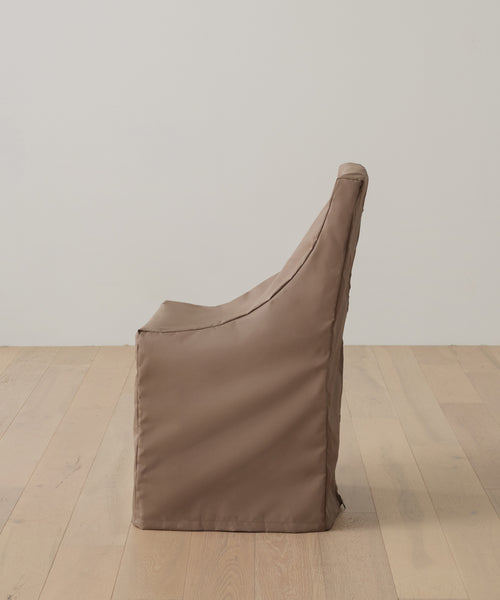 The Outdoor Montecito Dining Chair Cover in brown is displayed in profile on a light wooden floor, set against a plain off-white wall.