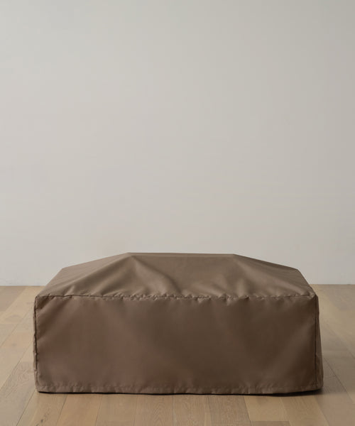 The Outdoor Vista Coffee Table Cover, in rectangular brown fabric, rests on a light wood floor against a plain off-white wall, resembling an indoor ottoman.