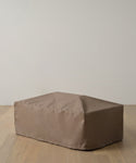 The Outdoor Vista Coffee Table Cover, a rectangular taupe fabric piece, rests on a light wooden floor against an off-white wall, offering a stylish look for your indoor or outdoor space.