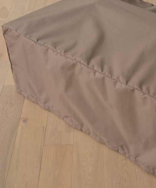 A close-up of the Outdoor Vista Coffee Table Cover, a light brown, rectangular fabric cover resembling an outdoor furniture accessory, placed on a light wood floor.