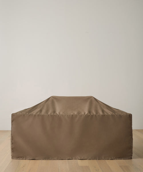 The Outdoor Vista Dining Table Cover, made of durable brown polyester, protects a large rectangular dining table placed on a light wood floor beside a plain white wall.