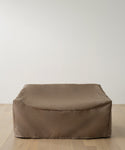 A large, rectangular brown bean bag sofa sits on a light wooden floor against a plain white wall, protected by the Outdoor Vista Sofa Cover for minimalist and modern patio furniture waterproof protection.