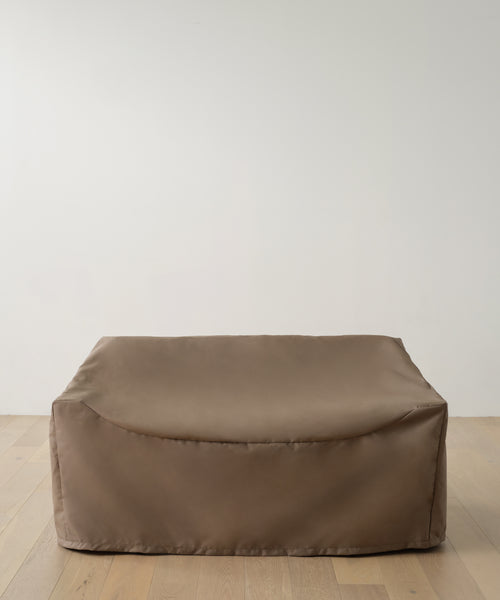 A large, rectangular brown bean bag sofa sits on a light wooden floor against a plain white wall, protected by the Outdoor Vista Sofa Cover for minimalist and modern patio furniture waterproof protection.