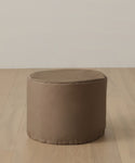 A round, light brown ottoman with a smooth surface sits on a light wood floor by an off-white wall—pair it with the Outdoor Marin Low Side Table Cover for enhanced protection and style.