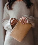 A woman in a beige knit sweater holds the Pony Hair Zip Pouch, a tan leather clutch handmade in Italy, with both hands and partially unzips it; only her torso and hands are visible in the cropped image.