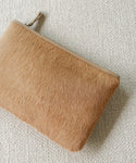 The Pony Hair Zip Pouch, a tan rectangular leather clutch with a furry texture and zipper closure, handmade in Italy, rests on a textured light beige fabric surface.