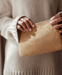 A person in a cream-colored sweater holds the Pony Hair Zip Pouch, a small, textured leather accessory handmade in Italy, with both hands against a soft, neutral background.