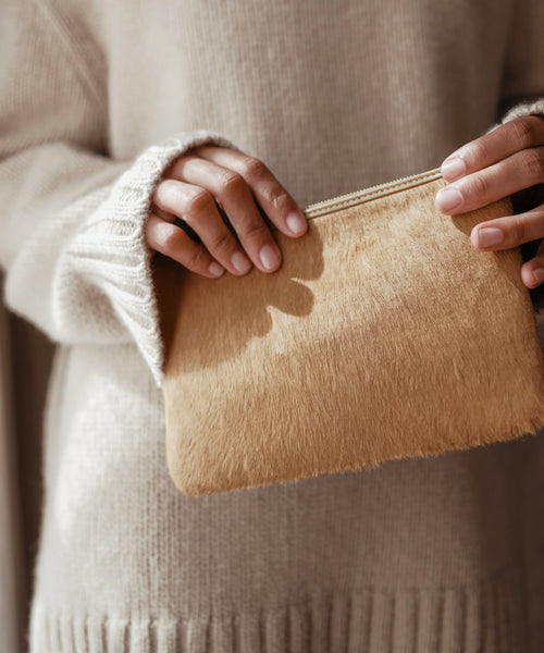 A person in a cream-colored sweater holds the Pony Hair Zip Pouch, a small, textured leather accessory handmade in Italy, with both hands against a soft, neutral background.