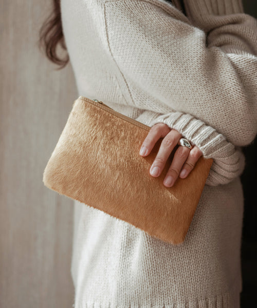 A person in a cream knit sweater holds the Pony Hair Zip Pouch, a tan furry leather clutch handmade in Italy, under their arm; only their torso and ring-adorned hand are visible.