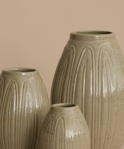 Three ribbed ceramic vases in a speckled light beige glaze, arranged against a solid warm beige background.