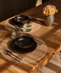 A rustic dining table set with two place settings featuring dark brown ceramic bowls, matching plates, clear glass tumblers, woven round placemats, silver cutlery with white handles, and a ribbed beige vase holding yellow-orange flowers.