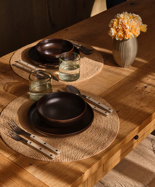 A rustic dining table set with two place settings featuring dark brown ceramic bowls, matching plates, clear glass tumblers, woven round placemats, silver cutlery with white handles, and a ribbed beige vase holding yellow-orange flowers.