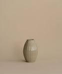 A small ribbed ceramic vase in a light warm gray-beige glaze, centered against a solid beige background.