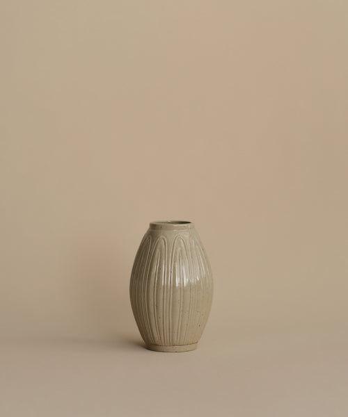 A small ribbed ceramic vase in a light warm gray-beige glaze, centered against a solid beige background.