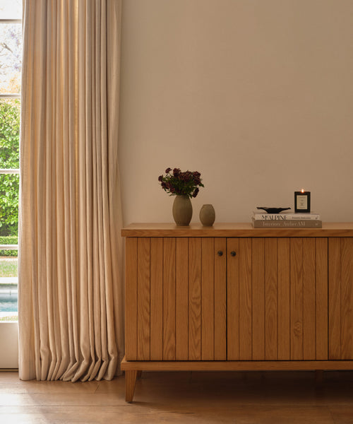 A wooden sideboard stands against a cream wall by a window with beige curtains. On it sits the Porta Ceramic Vase filled with flowers, stacked books, and a lit candle—perfect touches for elegant home décor as sunlight filters in.