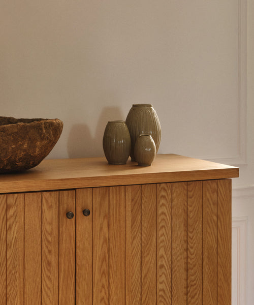 A wooden cabinet with vertical panels displays three Porta Ceramic Vases in muted tones and a rustic stone bowl, bringing timeless style to your home décor against a plain, light-colored wall.