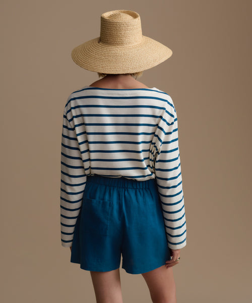 Person wearing a striped long-sleeve shirt and blue shorts with a woven hat on a beige background