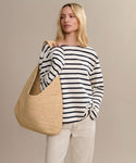 A woman stands against a plain beige background, carrying the Raffia Isla Bag while wearing a black and white striped long-sleeve shirt and light beige pants.