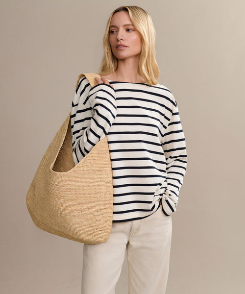 A woman stands against a plain beige background, carrying the Raffia Isla Bag while wearing a black and white striped long-sleeve shirt and light beige pants.